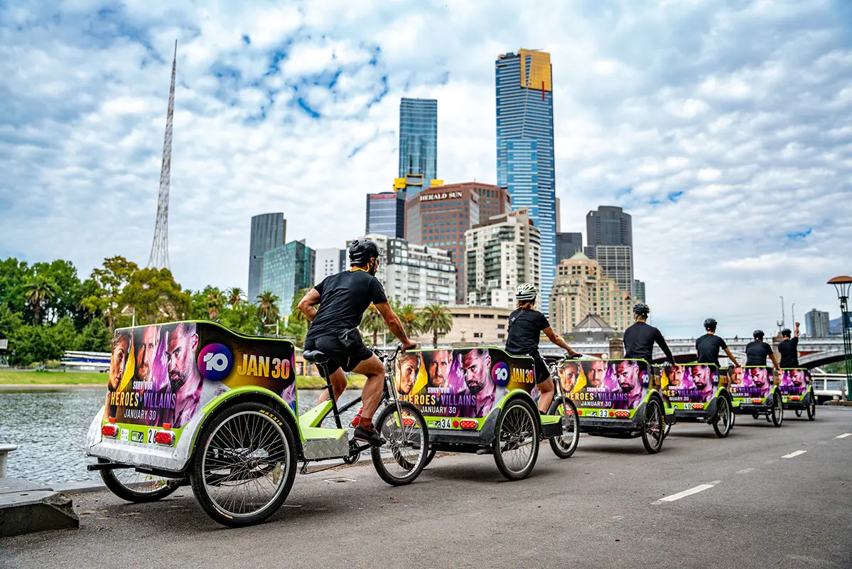 A line of pedicabs carrying television advertising panels riding along the Yarra with the Melbourne skyline behind.