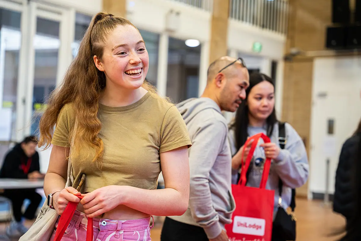A smiling student holding a lanyard at a university open-day stall, other visitors behind her.