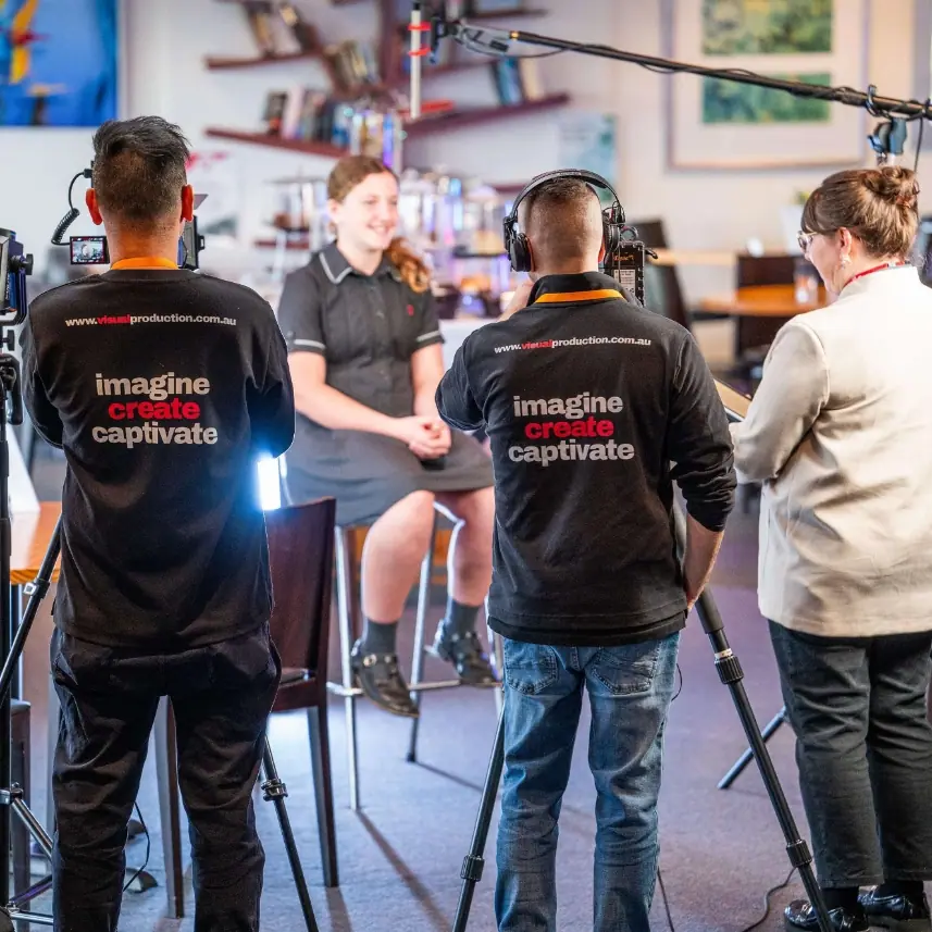 Two Visual Production Agency crew in “imagine create captivate” branded shirts filming a seated interview on location, one operating camera and one on sound.