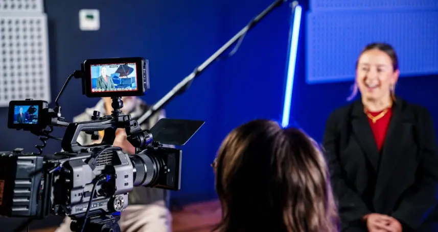 A cinema camera and boom microphone set up to film a woman being interviewed against a blue-lit studio backdrop.