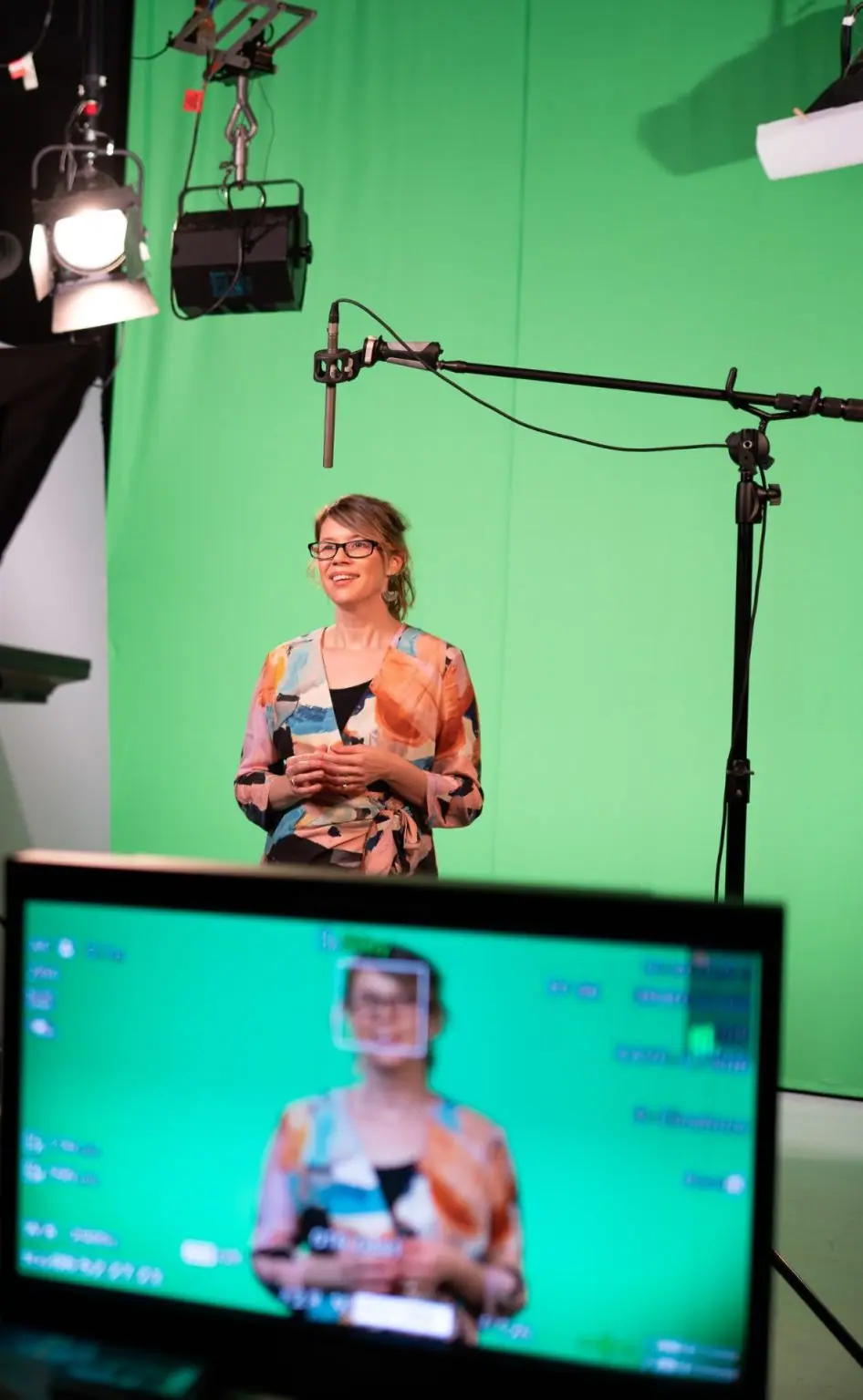 A presenter being filmed against a green screen in the Visual Production Agency studio, with studio lighting, an overhead boom microphone and a monitor showing the live camera feed.